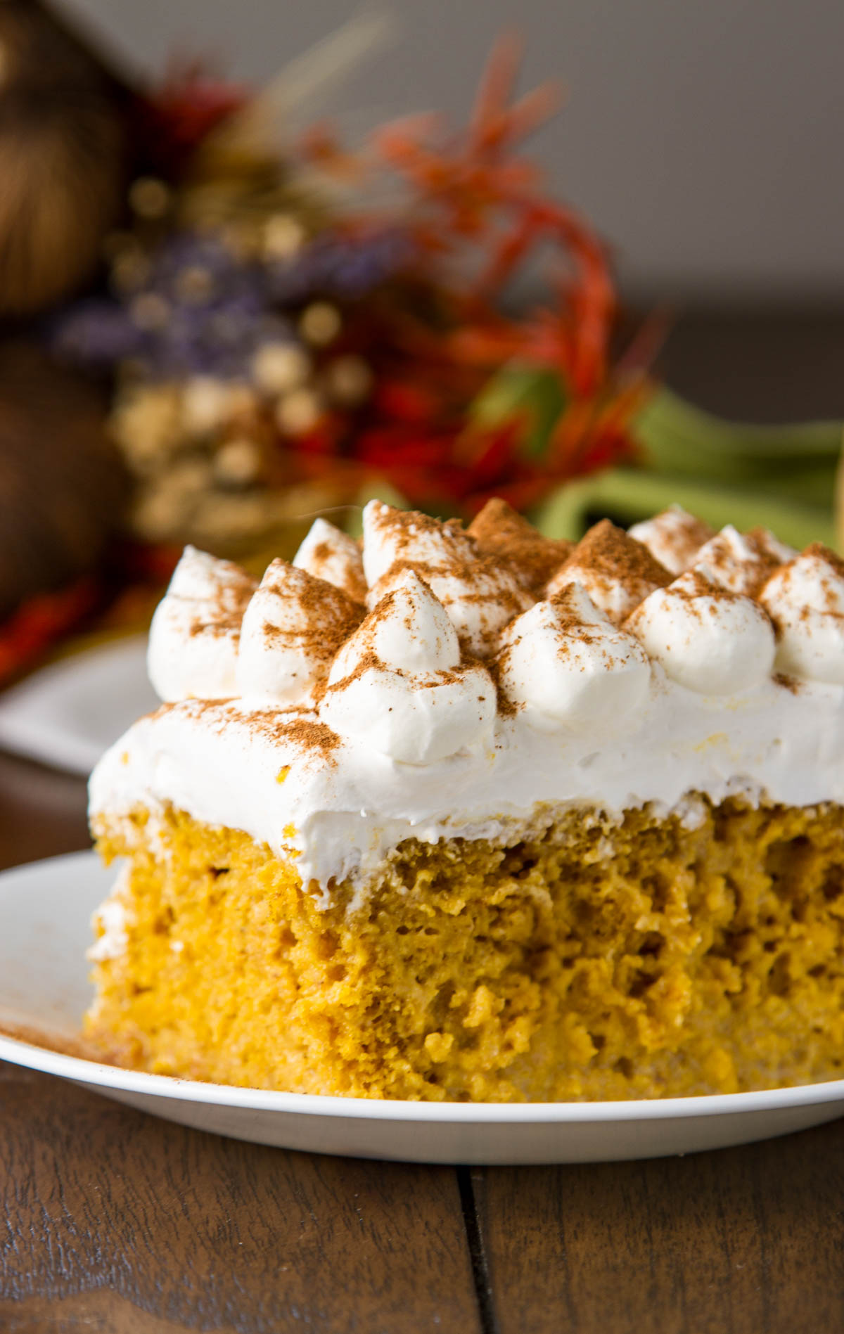 a slice of pumpkin tres leches cake on a small white plate on a wood table with fall decorations in the background