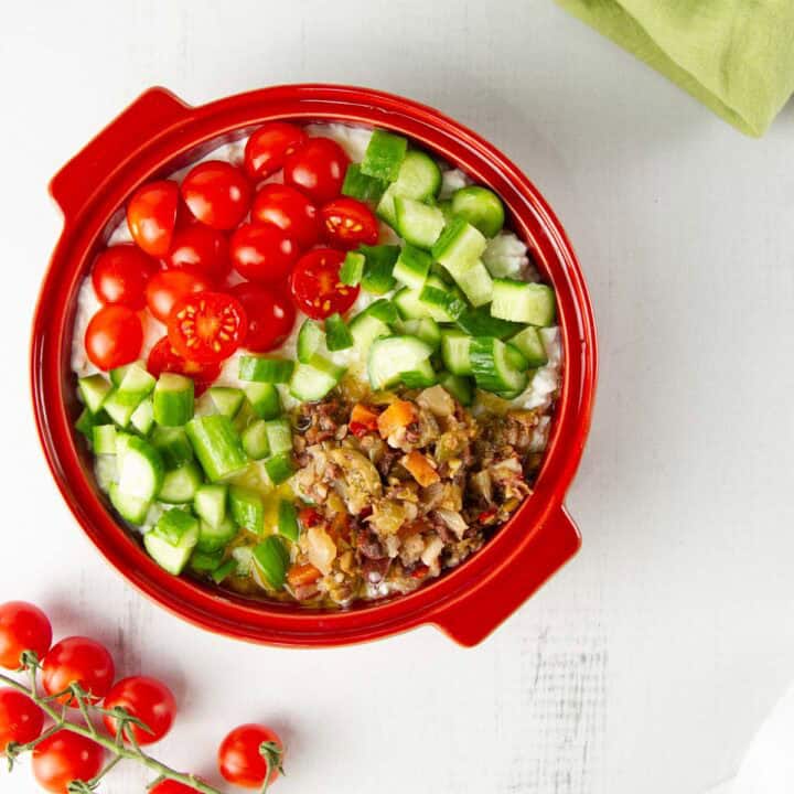 a mediterranean cottage cheese bowl in a round, red bowl on a white table next to fresh tomatoes on the vine