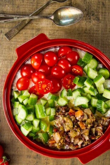a mediterranean cottage cheese bowl in a round, red bowl on a wood table next to fresh tomatoes on the vine and two spoons