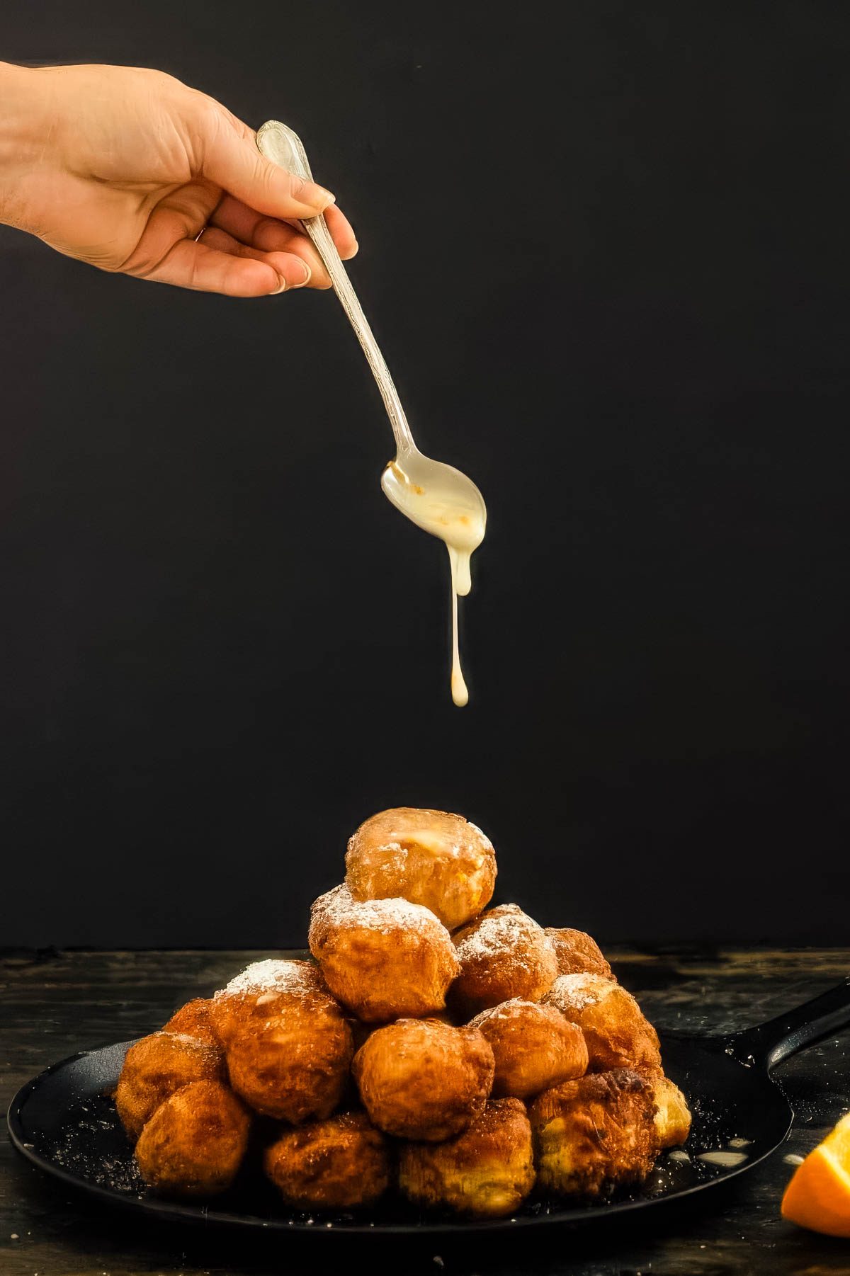 a pile of glazed donut holes on a black plate being drizzled with an orange glaze
