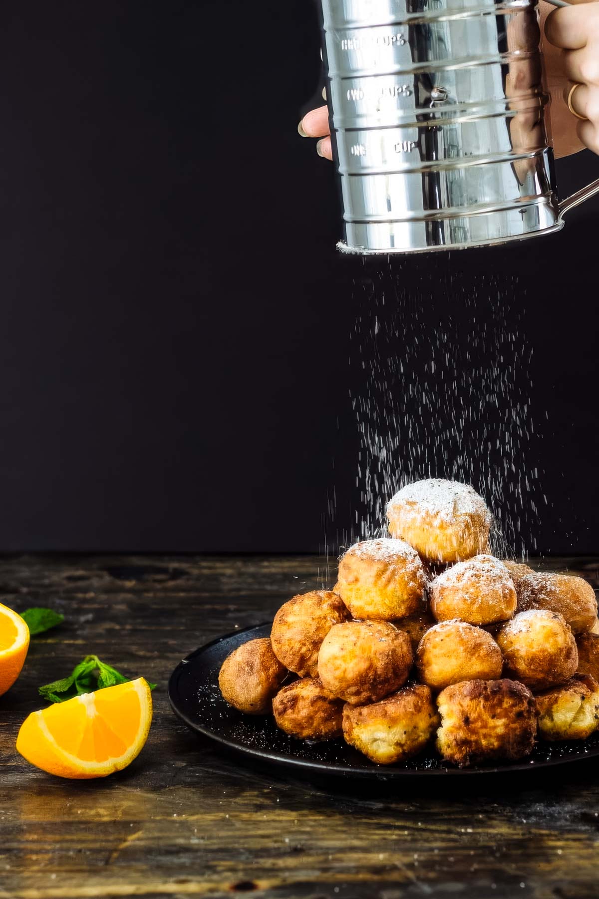 a pile of glazed donut holes on a black plate being sprinkled with powdered sugar next to two fresh orange wedges