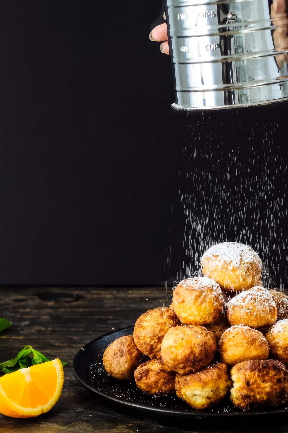 a pile of homemade donut holes on a black plate being sprinkled with powdered sugar next to two fresh orange wedges