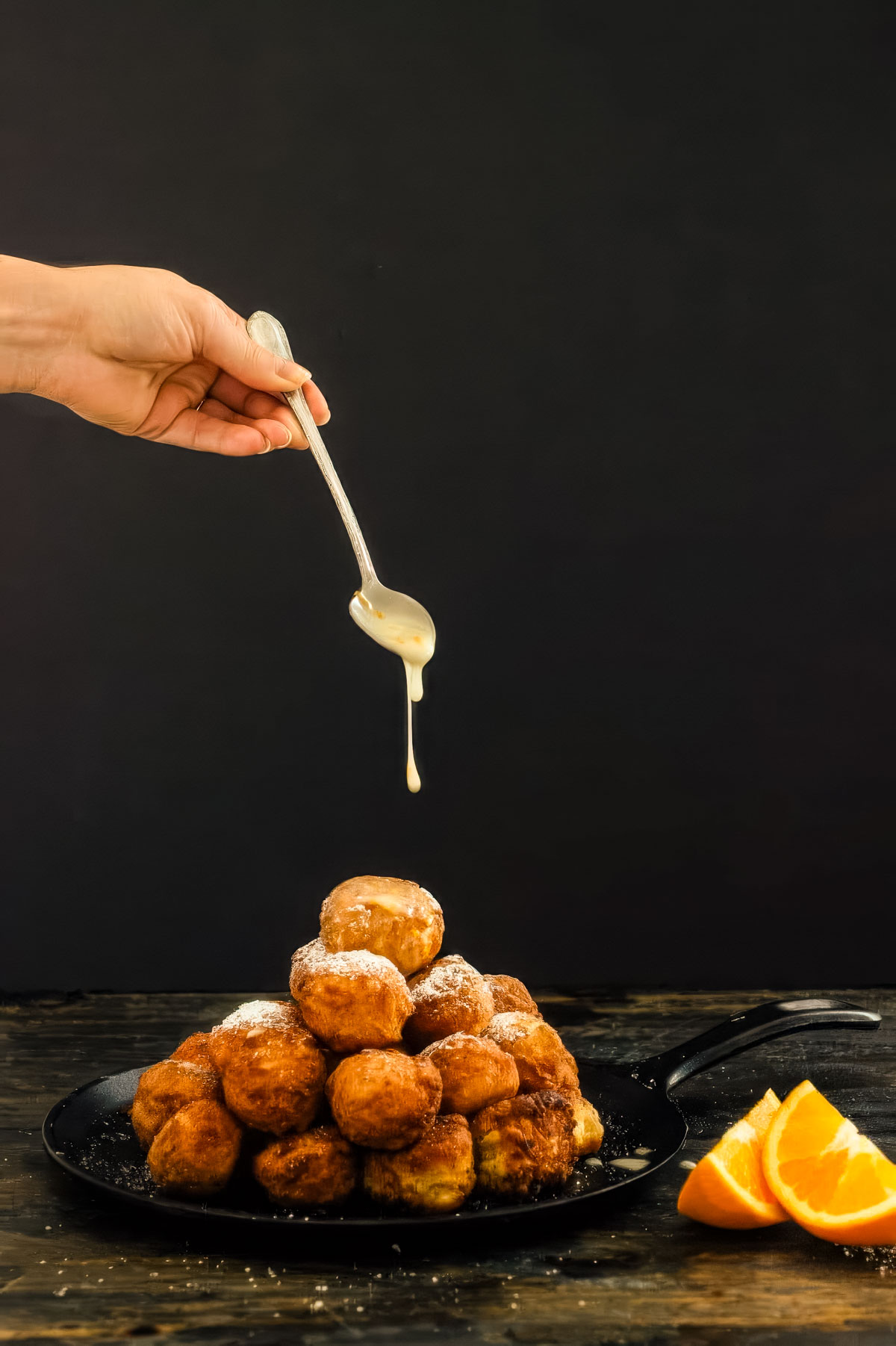 a pile of glazed donut holes on a black plate being drizzled with an orange glaze next to two fresh orange wedges