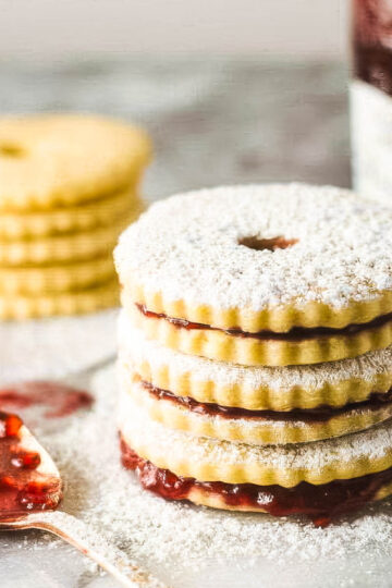 a stack of three jam sandwich cookies dusted with powdered sugar, next to cookies ready to be filled, along with a jar of raspberry jam and a spoon covered in jam