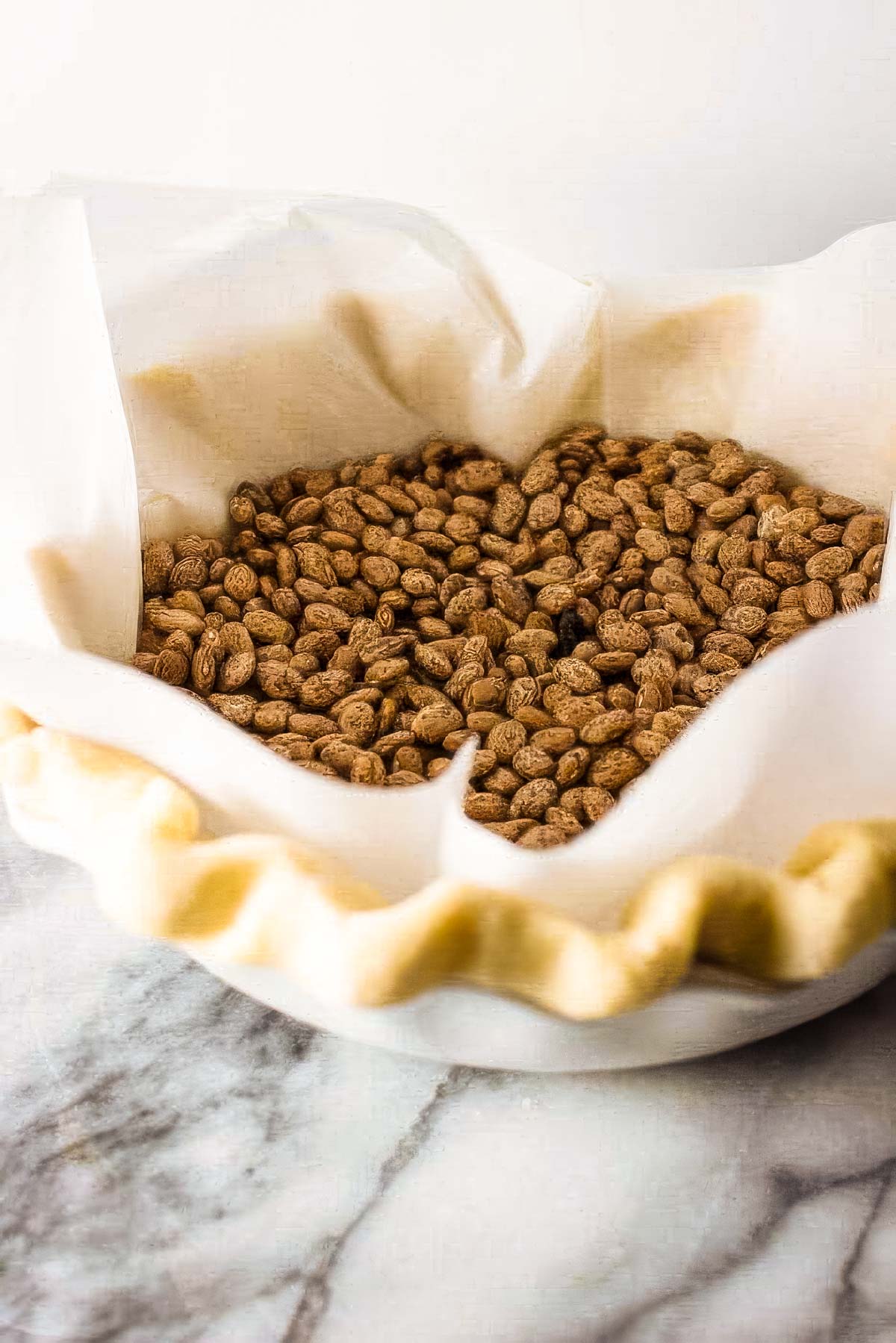 Step nine to making a homemade pie crust: a pie crust in a pie pan ready for the oven, with parchment paper then dried beans filling it for a blind bake