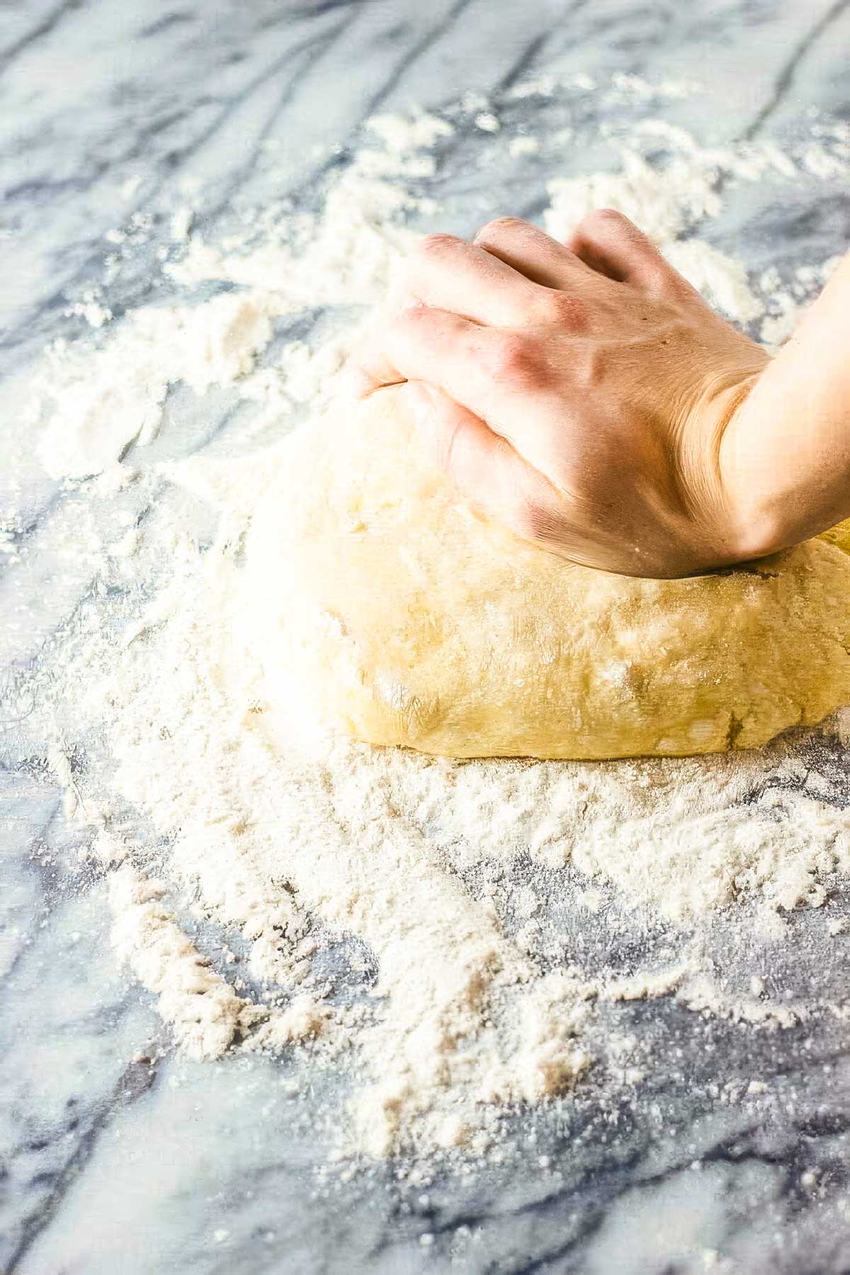 Step six to making a homemade pie crust: a hand pressing into the dough on a floured surface in preparation for rolling it out