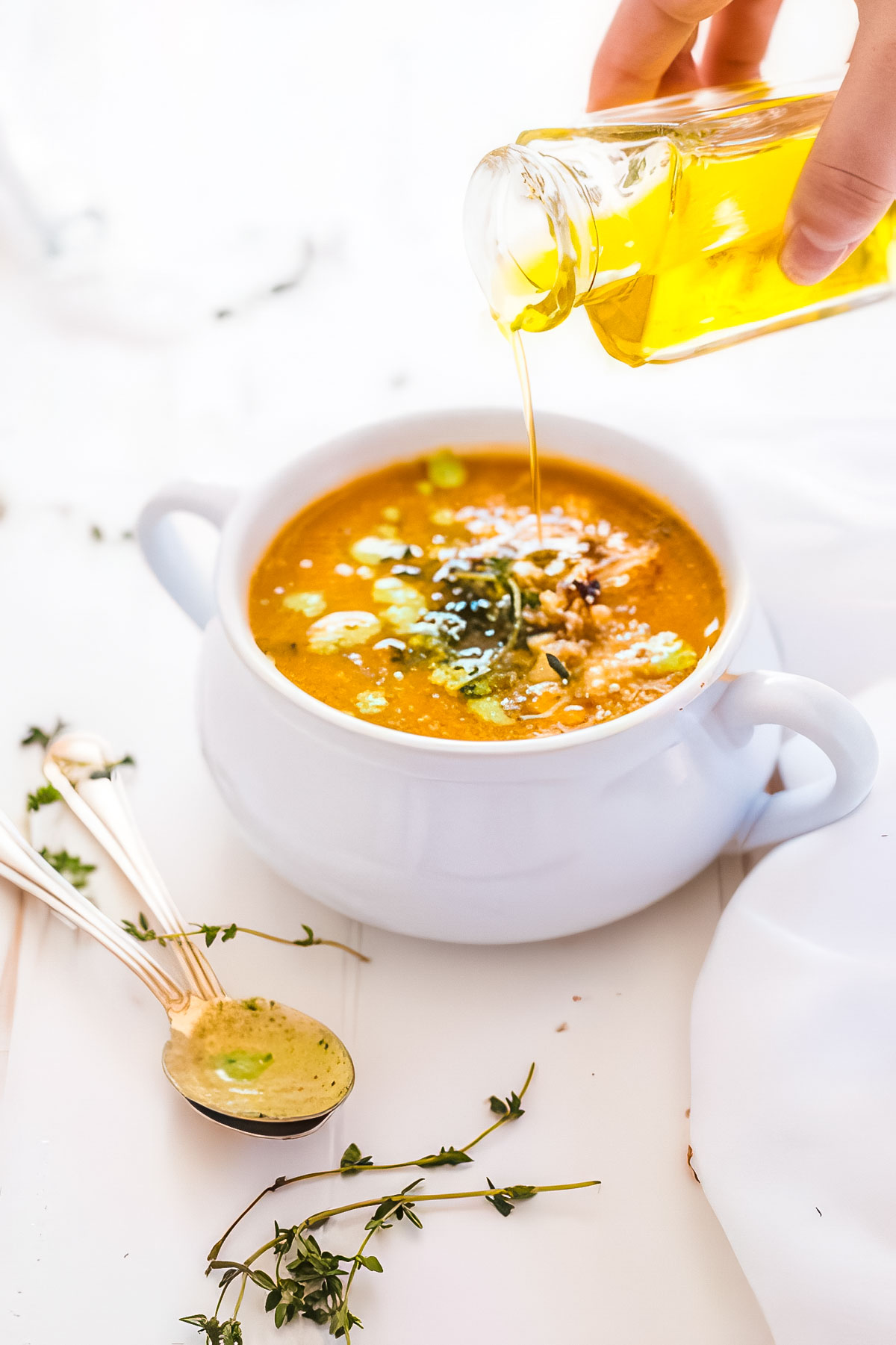 Roasted Cauliflower Tomato Soup with Parsley Cream a white bowl filled with cauliflower tomato soup with olive oil from a small jar being drizzled over the top next to two vintage spoons and fresh sprigs of thyme