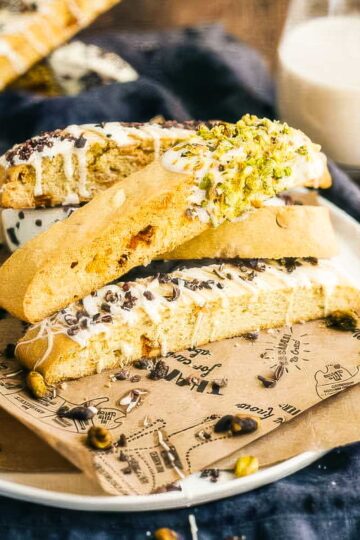 a plate of white chocolate biscotti, some coated with chopped pistachios and some with chopped cacao nibs, next to a glass of milk