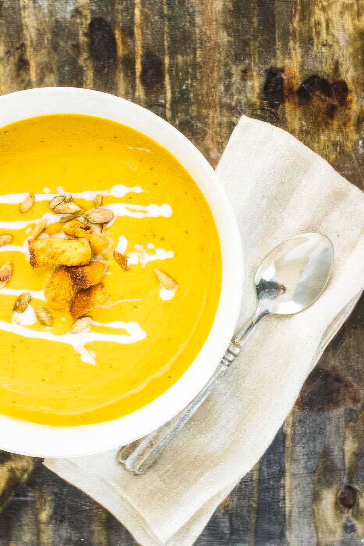 an overhead view of a white bowl filled with butternut squash and sweet potato soup, next to a white napkin and a spoon an a rustic wood table