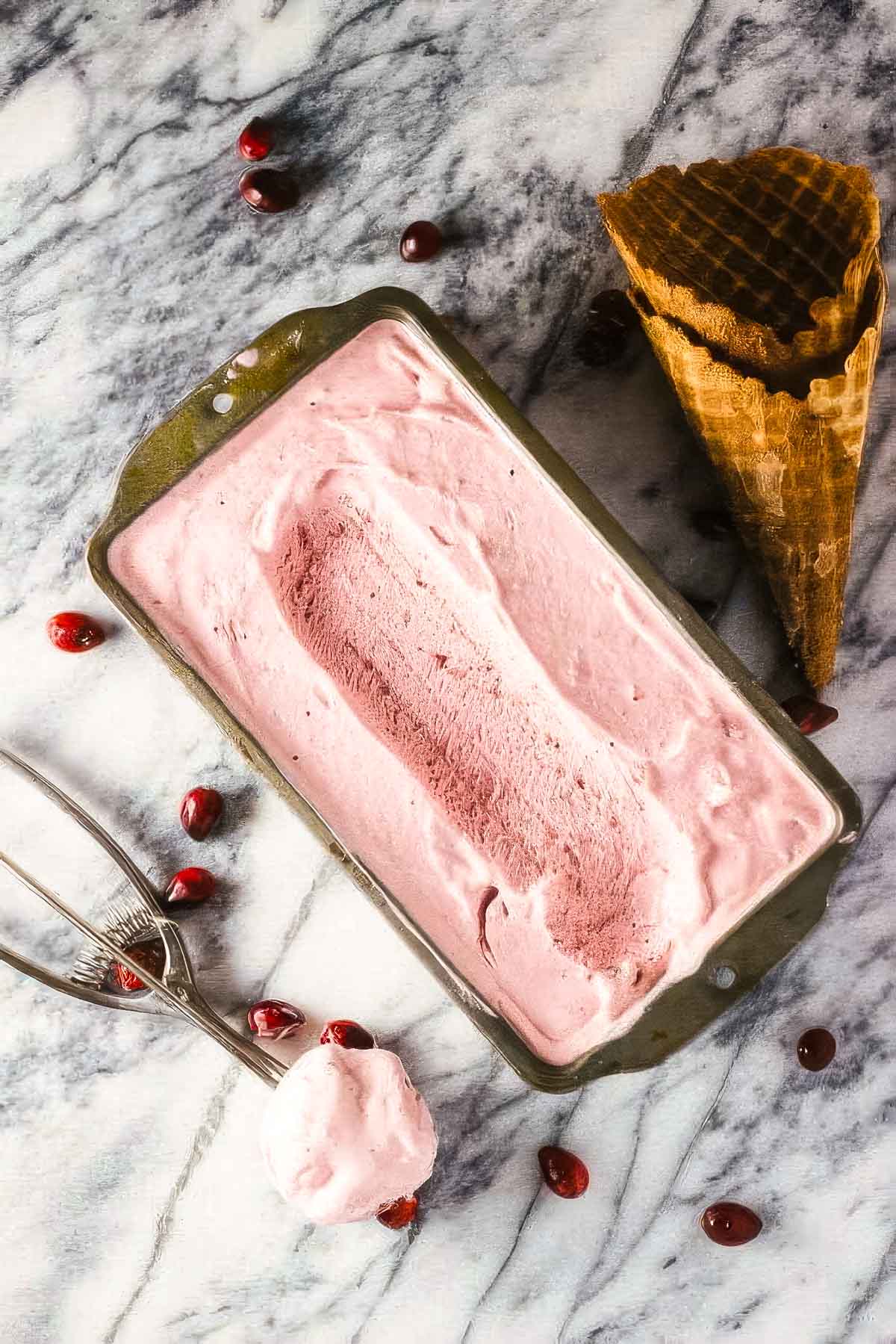 a vintage loaf pan filled with cranberry ice cream on a marble countertop with an ice cream scoop, two waffle cones, and a few fresh cranberries scattered around it