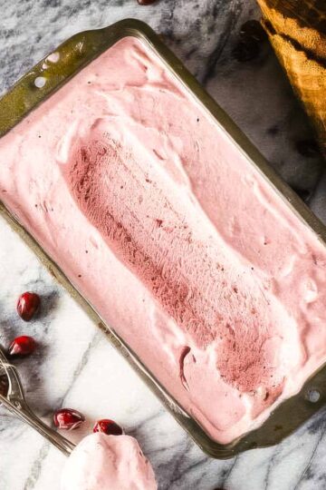 a vintage loaf pan filled with cranberry ice cream on a marble countertop with an ice cream scoop, two waffle cones, and a few fresh cranberries scattered around it