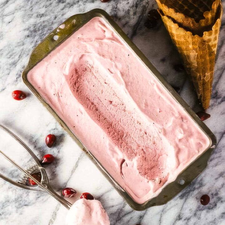 a vintage loaf pan filled with cranberry ice cream on a marble countertop with an ice cream scoop, two waffle cones, and a few fresh cranberries scattered around it