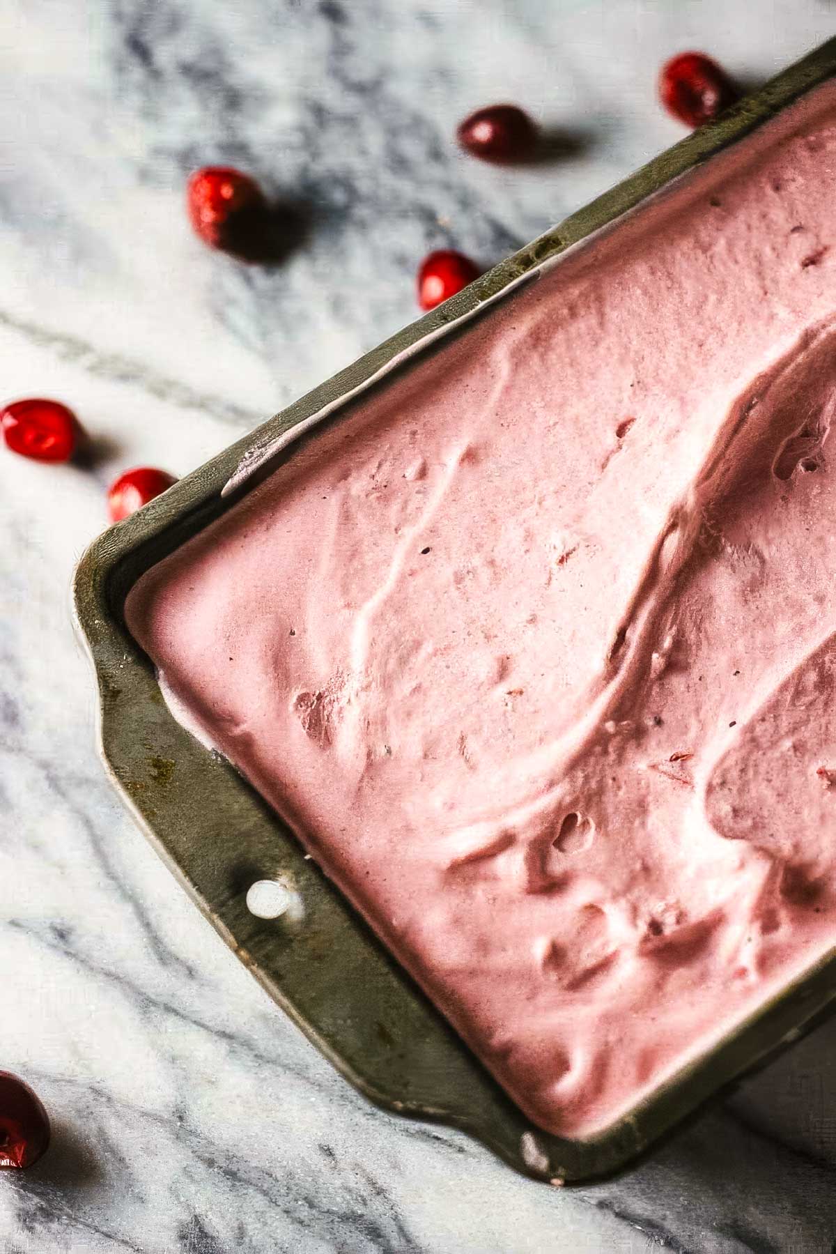 a vintage loaf pan filled with cranberry ice cream on a marble countertop with a few fresh cranberries scattered around it