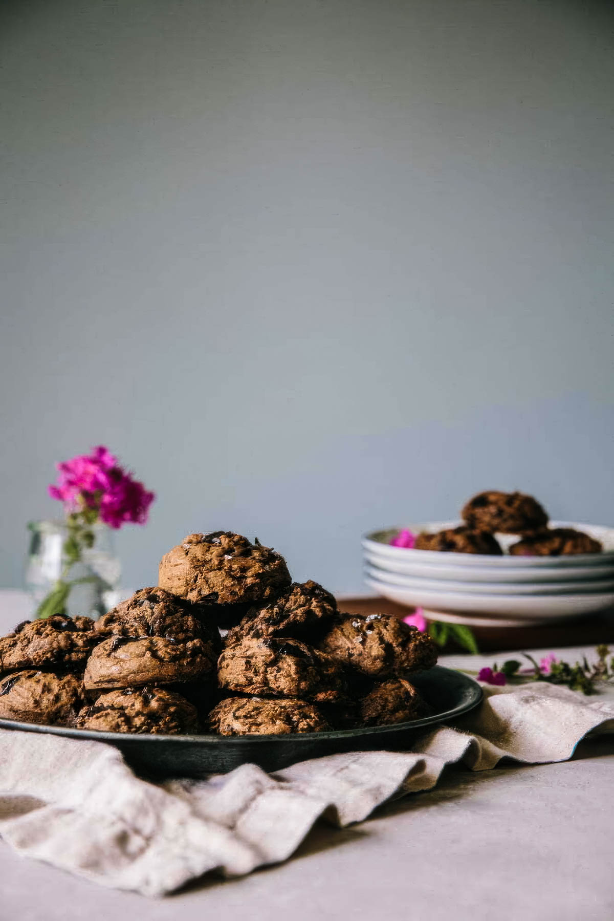 a side view of a vintage plate filled with double chocolate cookies topped with sea salt next to a pink flower in a vase
