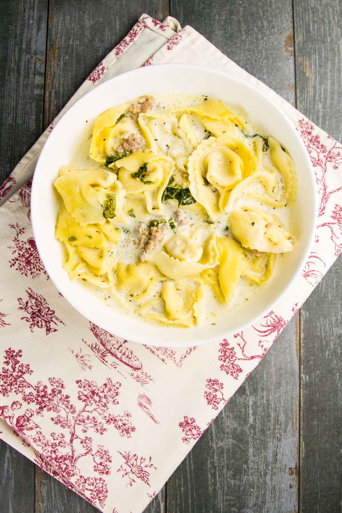 Sausage Tortellini Soup (Freezer Friendly) a bowl of sausage tortellini soup with kale on a vintage towel on a gray wood table