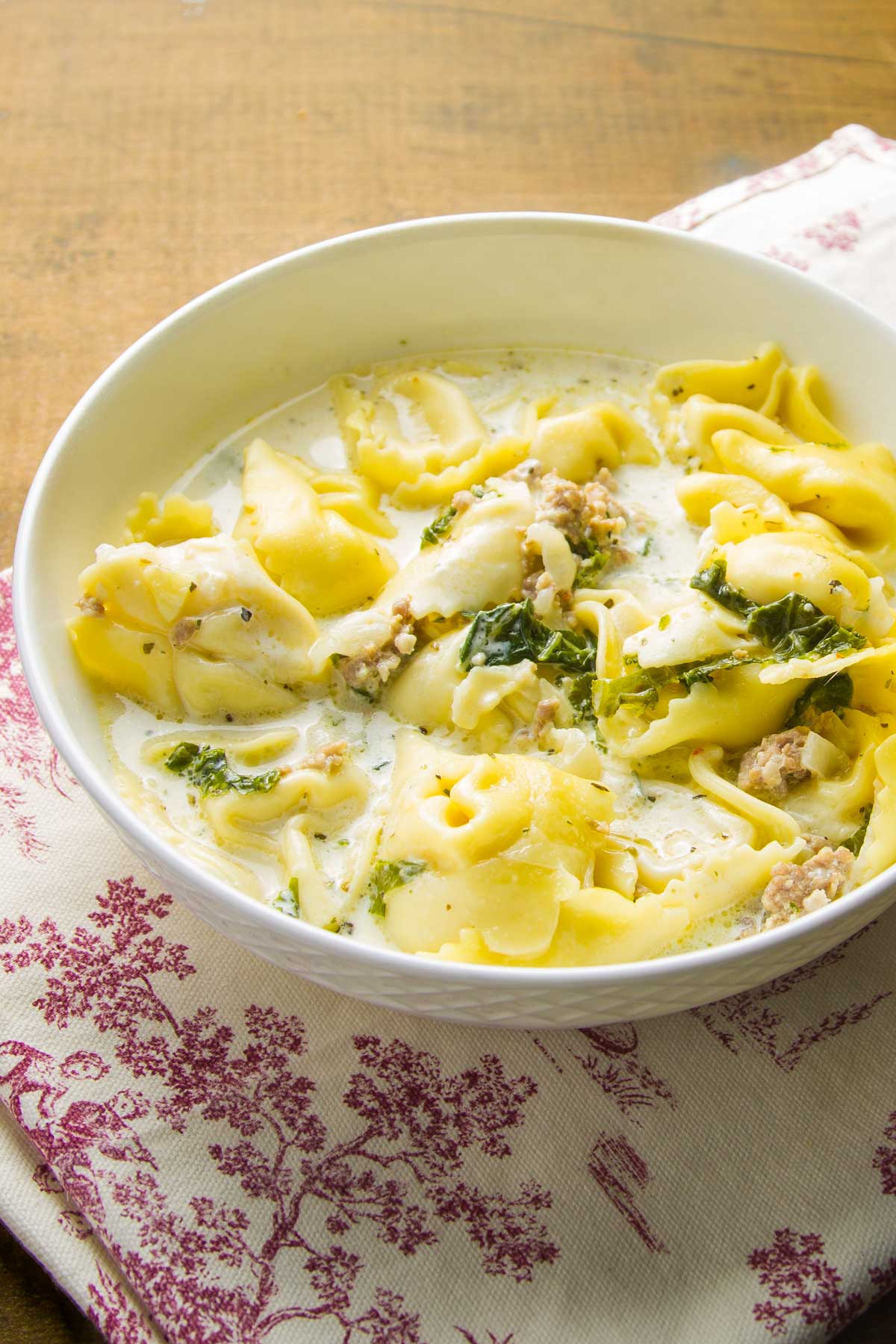 Sausage Tortellini Soup (Freezer Friendly) a side view of a bowl of sausage tortellini soup with kale on a vintage towel on a wood table