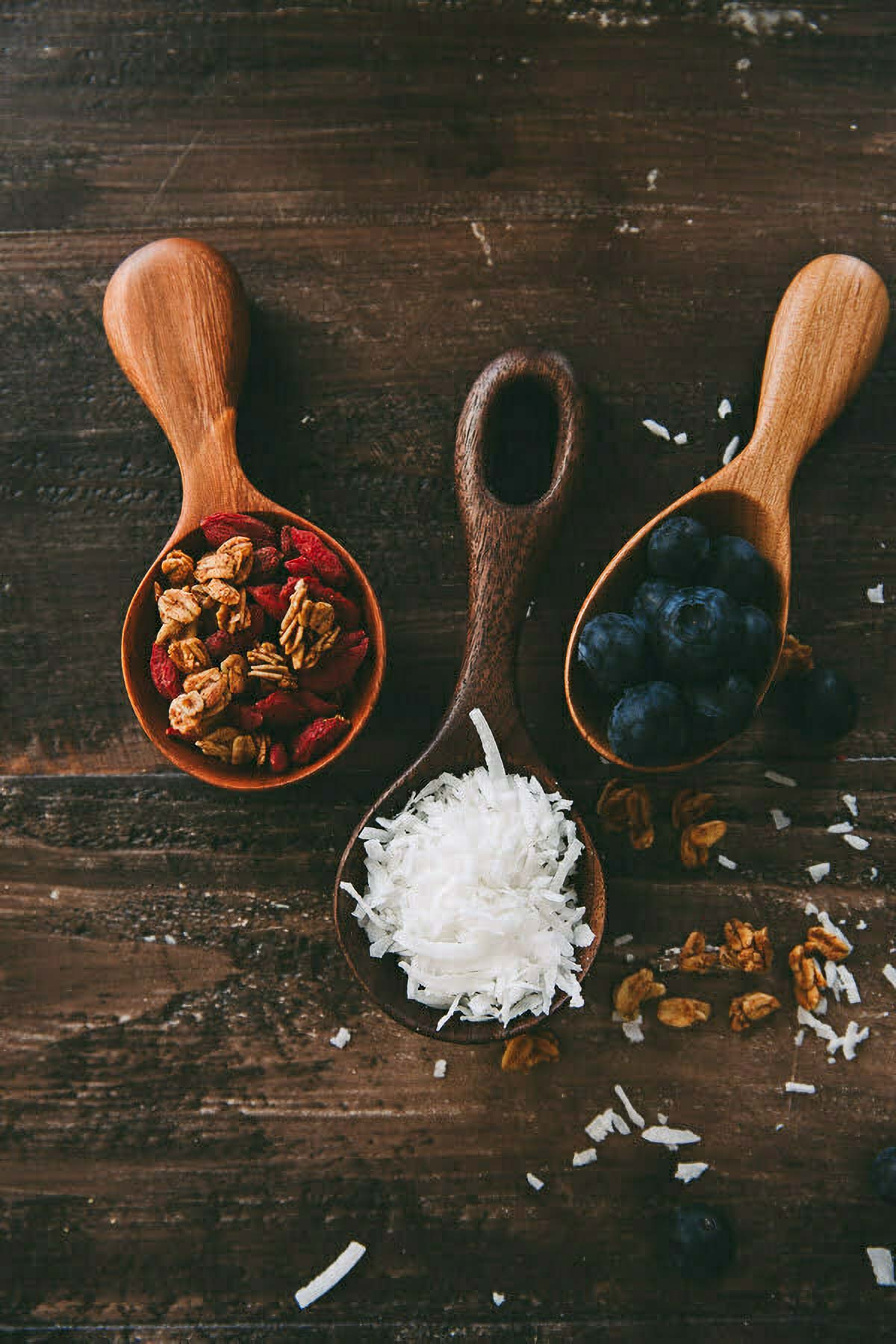 Coconut Smoothie Bowl three wooden scoops filled with toppings for a coconut smoothie bowl (granola and goji berries, fresh blueberries, and shredded coconut)