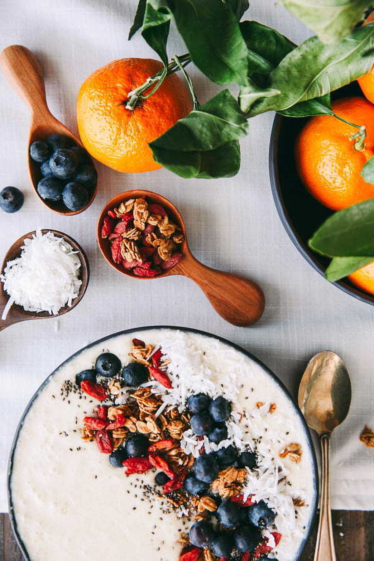 Coconut Smoothie Bowl a coconut smoothie bowl topped with fresh blueberries and granola next to wooden scoops filled with the toppings and a bowl of fresh oranges