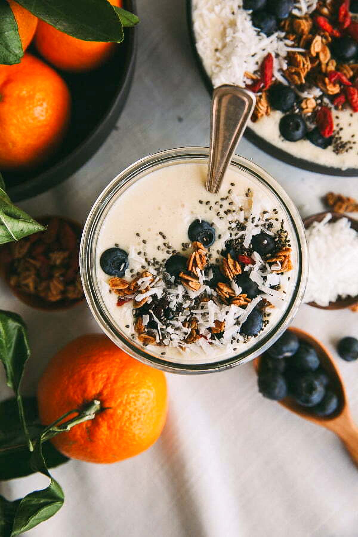 Coconut Smoothie Bowl a coconut smoothie bowl topped with fresh blueberries and granola next to wooden scoops filled with the toppings and a bowl of fresh oranges