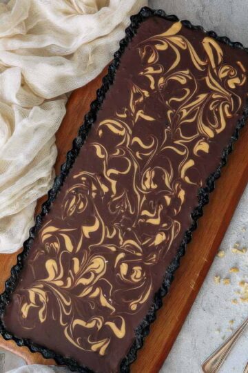 Chocolate Ganache Tart with Peanut Butter Swirl (No Bake!) a top-down view of a chocolate ganache tart with a decorative peanut butter swirl on an angle next to a beige towel and two spoons with crushed peanuts