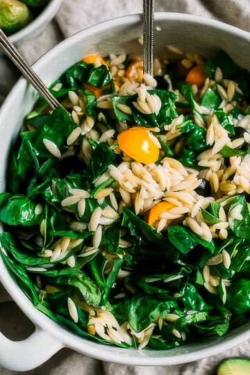 a large white bowl filled with orzo spinach salad and halved yellow cherry tomatoes on a light tan linen towel