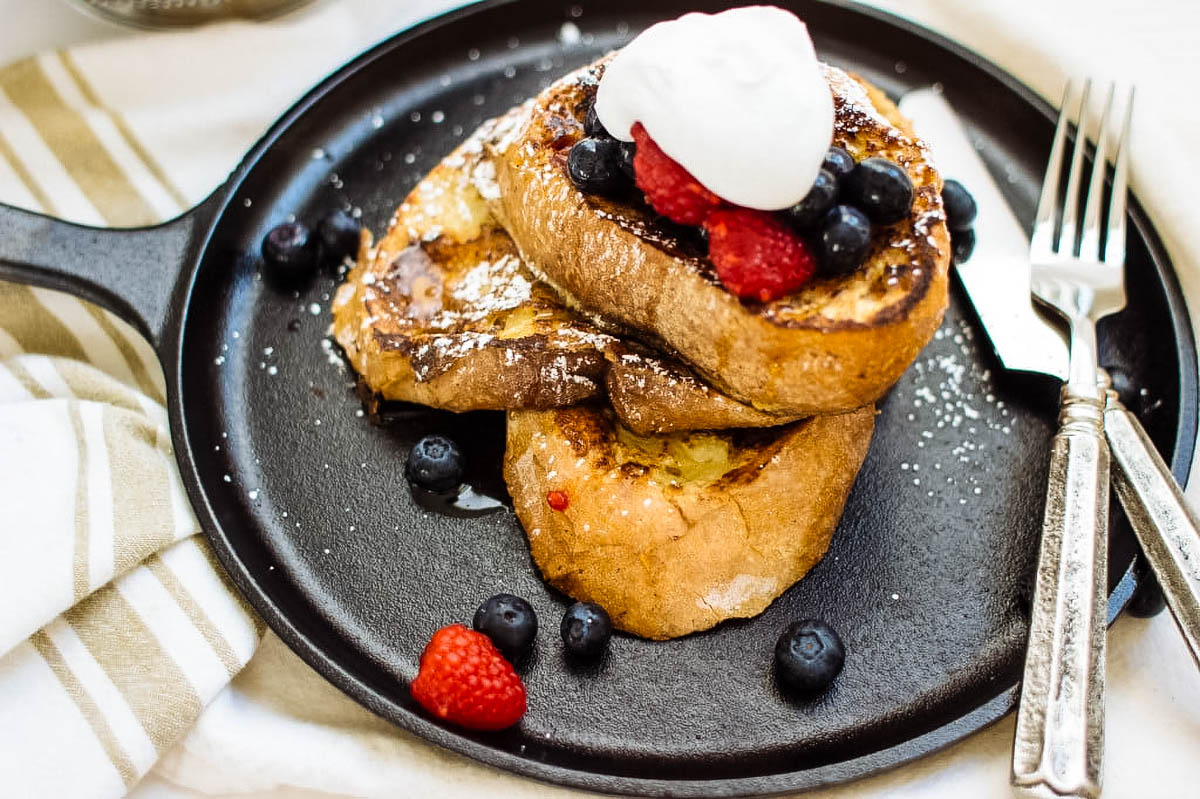 a stack of sourdough french toast on a black plate topped with fresh berries and mascarpone cream