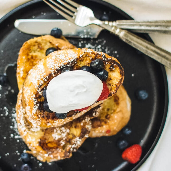 a stack of sourdough french toast on a black plate topped with fresh berries and mascarpone cream