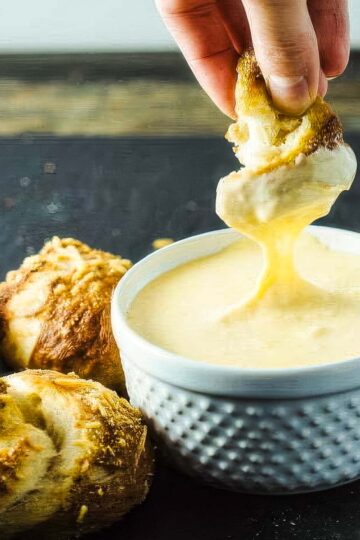 several garlic pretzel knots next to a dish of warm cheese sauce with a person dipping