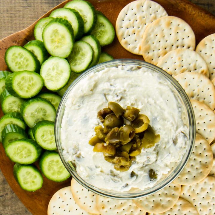 a closeup of a bowl of cream cheese olive dip topped with chopped green olives on a round wooden board with water crackers and sliced Persian cucumbers