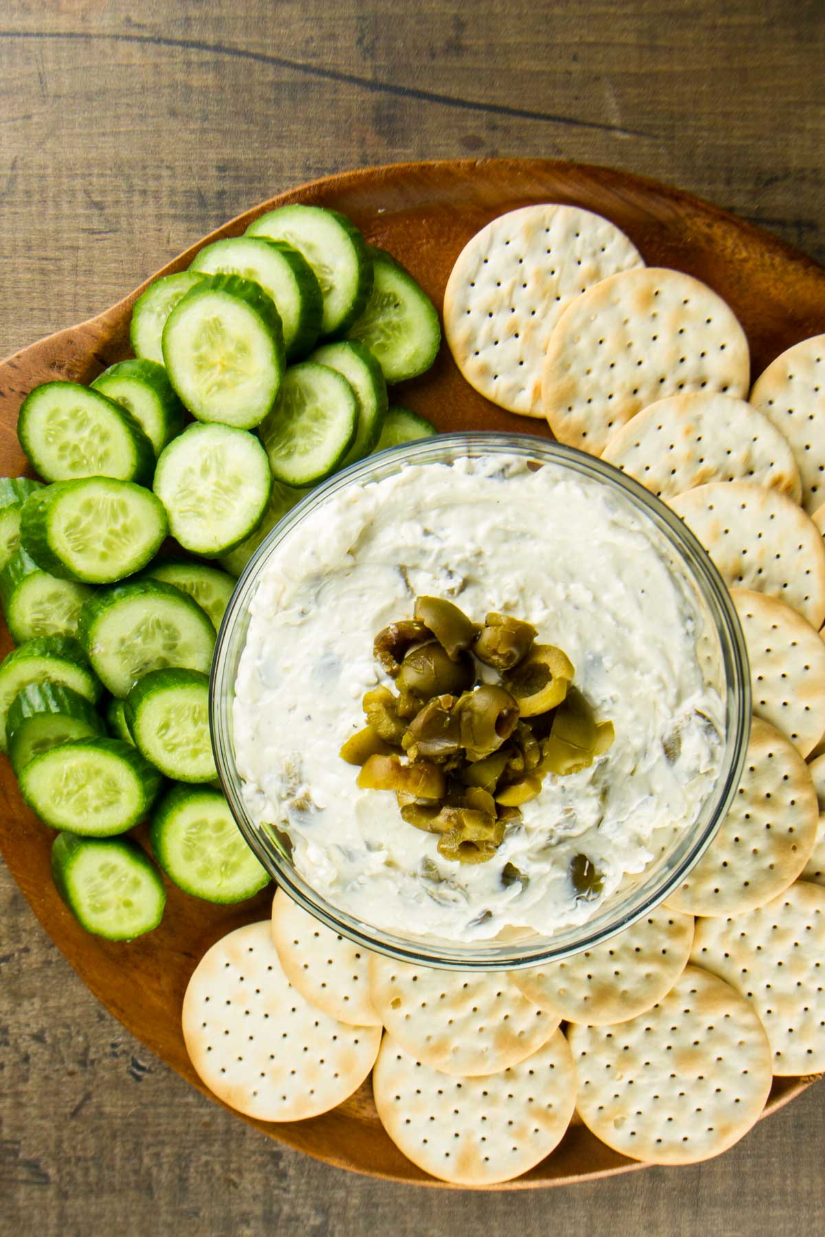 a closeup of a bowl of cream cheese olive dip topped with chopped green olives on a round wooden board with water crackers and sliced Persian cucumbers