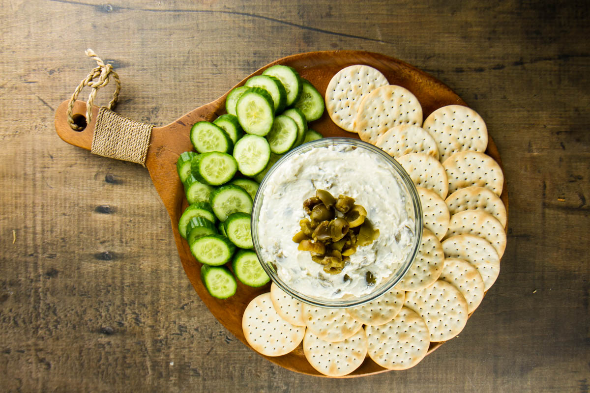 a bowl of cream cheese olive dip topped with chopped green olives on a round wooden board with water crackers and sliced Persian cucumbers